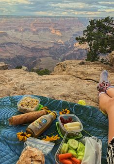 a woman sitting on top of a blue blanket next to food and drinks at the edge of a cliff