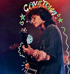 a person with headphones on holding a guitar in front of a neon sign that says cavetown