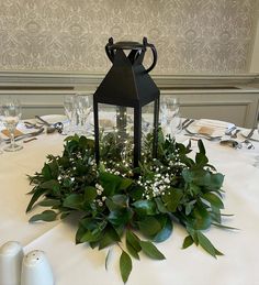 a black lantern on top of a table surrounded by greenery and white candlesticks