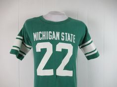 "Vintage 1970s or 80s college t shirt. Made of green cotton with thick white college graphics: Michigan State #22. Has Champion blue block label. Size large. Actual measurements are: 39\" around the chest 39\" around the waist 20\" shoulder seam to shoulder seam 28\" overall length In very good condition with 3\" of seam repair to the cuff." Retro College T-shirt With Screen Print, Retro College T-shirt For Football Season, Vintage Style Screen Print Fan Gear T-shirt, Vintage Screen Print T-shirt For Fans, Vintage Screen Print Fan Gear T-shirt, College Team Name Green T-shirt, Vintage Graphic Print Football T-shirt, Vintage Graphic T-shirt For Football Season, Green College T-shirt With Team Name