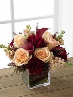 an arrangement of flowers in a square vase on a wooden table next to a window