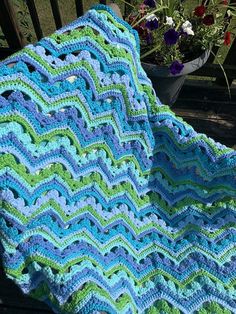 a blue and green crocheted blanket sitting on top of a wooden bench next to flowers