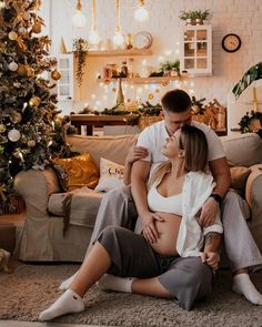 a pregnant couple sitting on the floor in front of a christmas tree