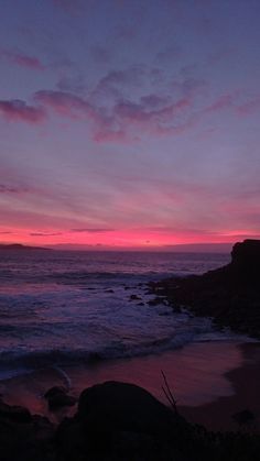 the sun is setting over the ocean with rocks in the foreground and pink clouds in the background