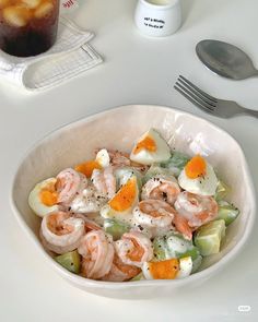 a white bowl filled with shrimp salad next to a fork and knife on a table