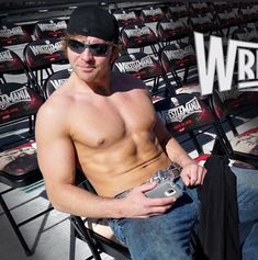 a shirtless man sitting in a stadium chair with his hand on his hip while holding a camera