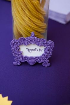 a purple table topped with a glass vase filled with yellow pastasticks next to a sign that reads raguel's hair