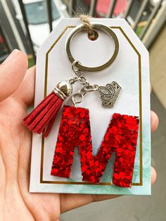 the letter m is made out of red glitter and has a tasseled keychain