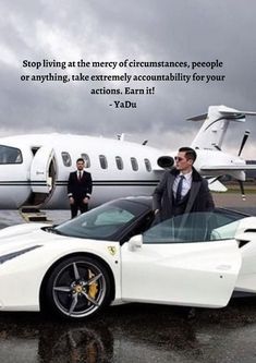 two men in suits standing next to a white sports car with an airplane behind them