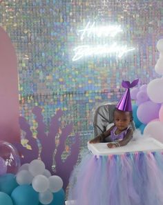 a baby sitting in a high chair wearing a purple and blue birthday hat with balloons around it