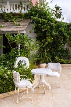 two white chairs and a table in front of some plants on the ground near a building