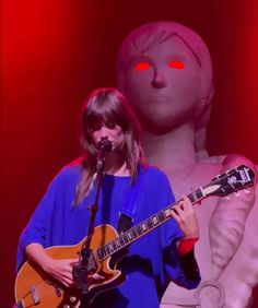 a woman is playing an electric guitar in front of a statue with glowing red eyes