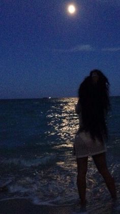 a woman standing on top of a beach next to the ocean under a full moon