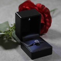 an engagement ring sits in a box next to a red rose on a tablecloth