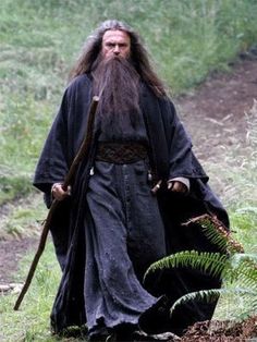 a man with long hair and beard walking in the woods holding a staff, wearing an old - fashioned black robe