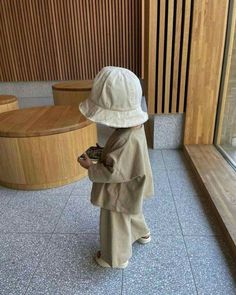 a little boy standing in front of a window wearing a white hat and trench coat