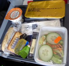 an airplane tray with some food and drinks on it, including bread, cheese, cucumbers, carrots, lettuce