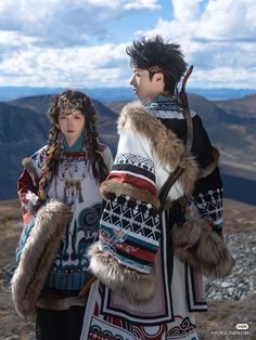 two people dressed in native clothing standing on top of a rocky hill with mountains in the background