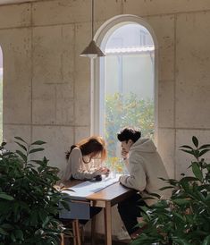 two people are sitting at a table looking out an open window, with plants in the foreground