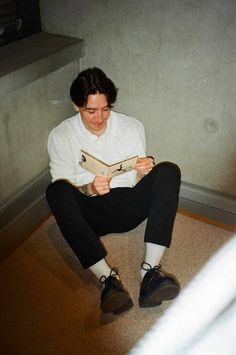 a young man sitting on the floor reading a book while wearing black pants and white shirt