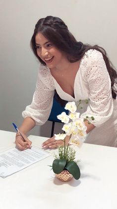 a woman sitting at a table signing a piece of paper with flowers in the vase