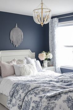 a white bed sitting under a chandelier next to a window with blue walls