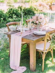 a wooden table topped with pink flowers and white plates next to a tall vase filled with greenery