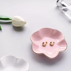 a pink flower shaped bowl with two small gold earrings on it next to a white tulip