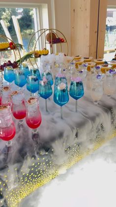 many glasses are lined up on a table in front of a window with colorful liquid