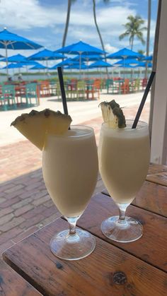 two glasses filled with drinks sitting on top of a wooden table next to an umbrella covered beach