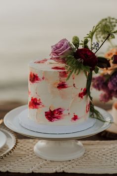 a white cake with red and pink flowers on it sitting on a table next to plates