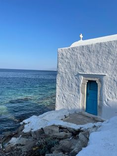 a white building with a blue door and cross on the roof next to the ocean