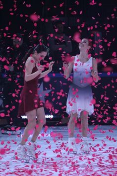 two women standing on a tennis court surrounded by confetti and pink petals in the air