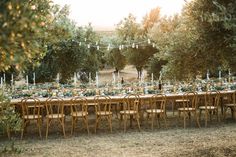 an outdoor dinner table set up with wooden chairs and tables covered in greenery, surrounded by trees