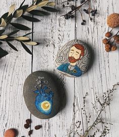 two painted rocks sitting on top of a white wooden table next to plants and nuts