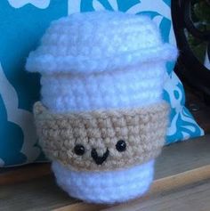 a crocheted coffee cup with a cute face on it's side sitting next to a pillow