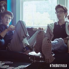 two young men sitting on a couch in front of a window with their feet up