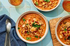 three bowls of pasta soup on a blue table with silverware and napkins next to it