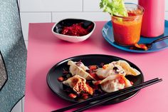 a pink table topped with black plates filled with food and chopsticks next to a drink