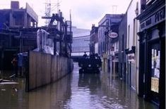 a flooded street with people walking on the sidewalk and cars parked in the alleys