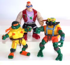 three toy action figures are posed on a white surface, one is wearing goggles and the other has an orange helmet