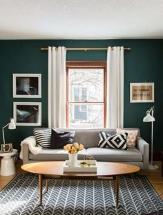 a living room with green walls and white drapes on the windowsill, a gray couch in front of a coffee table surrounded by black and white rugs