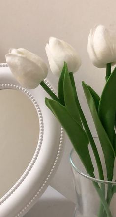 three white tulips in a clear glass vase next to a mirror on a table