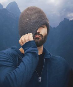 a man with a beard wearing a beanie and looking at the camera while standing in front of mountains