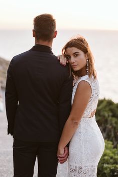 a man and woman standing next to each other in front of the ocean at sunset