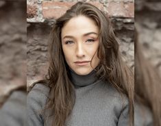 a woman with long hair standing in front of a brick wall wearing a turtle neck sweater