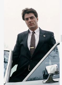 a man in a suit and tie leaning on the hood of a car with his hand out