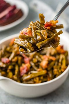 a spoon full of green beans and bacon in a white bowl with other dishes behind it