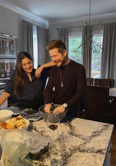 a man and woman sitting at a kitchen table with food in bowls on the counter