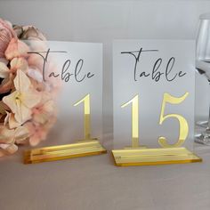 two place cards with gold numbers are on a table next to a wine glass and flowers